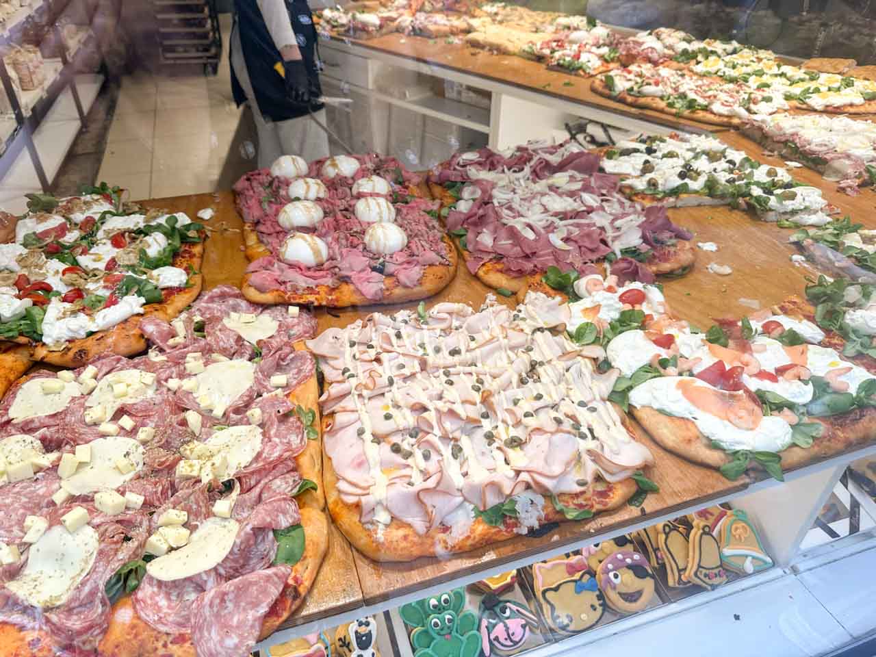 A variety of focaccia pizzas with different toppings such as cured meats, cheese, and fresh greens, laid out on a large wooden board at Il Fornaio in Bergamo, Italy